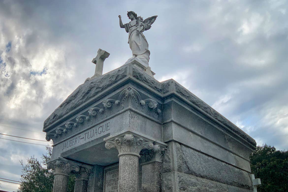 A grey stone mausoleum with a winged angel and cross on top in the St. Louis Cemetery No. 1.