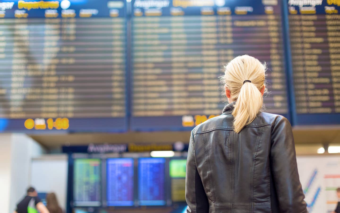 A blonde woman faces an airport Departure board in the terminal while wearing a black leather jacket.