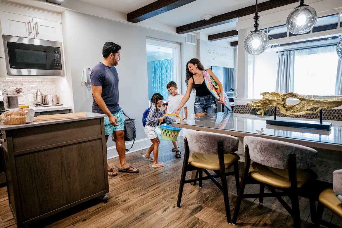 A man, woman, and young boy and girl wear and carry beach apparel and gear in the kitchen of a Signature Collection villa.