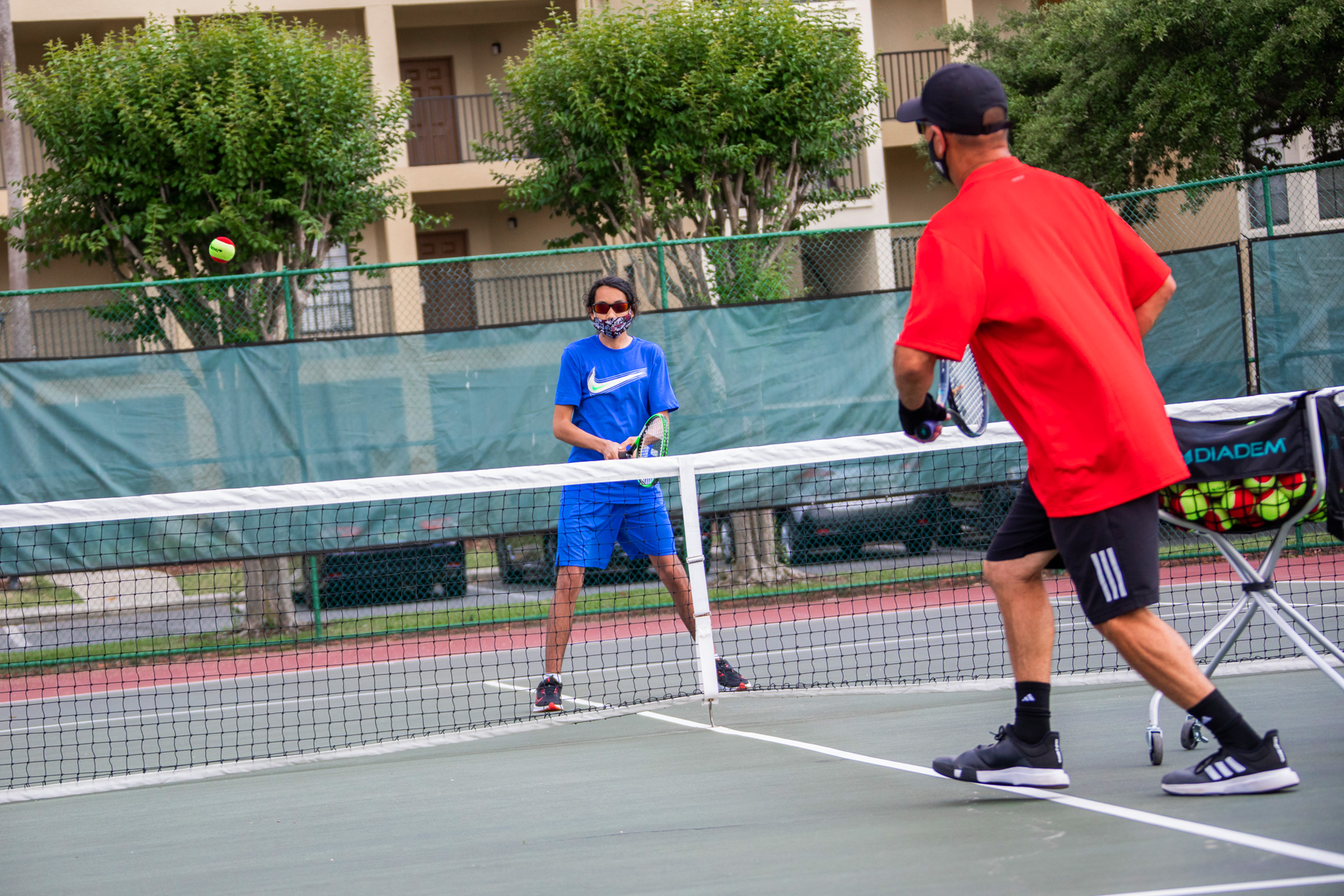 Special Olympic Athlete, Roan Luallen, plays tennis wearing a blue t-shirt and shorts with a safety mask and sunglasses with an instructor on the courts of our Orange Lake Resort located in Orlando, Florida.