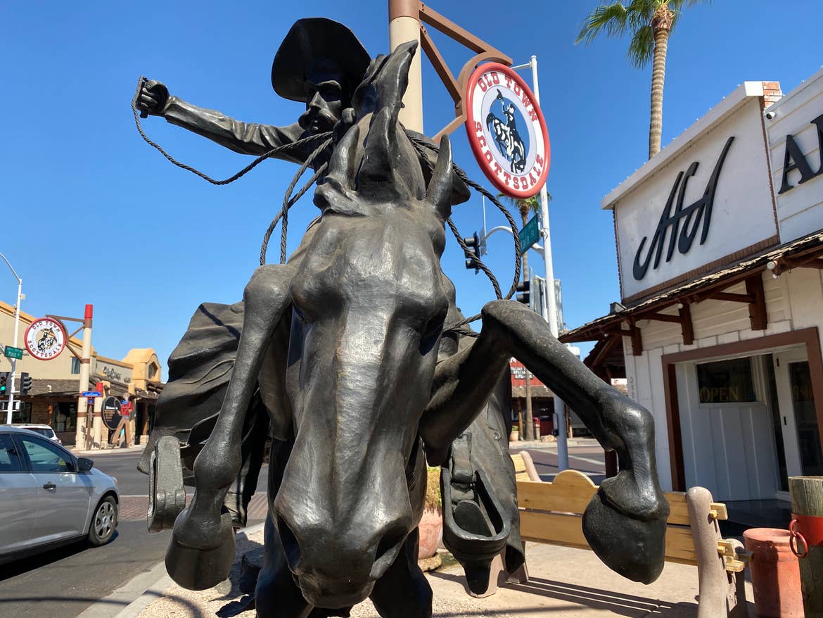 A cowboy statue in front of the Old Town Scottsdale sign with shops in the background.