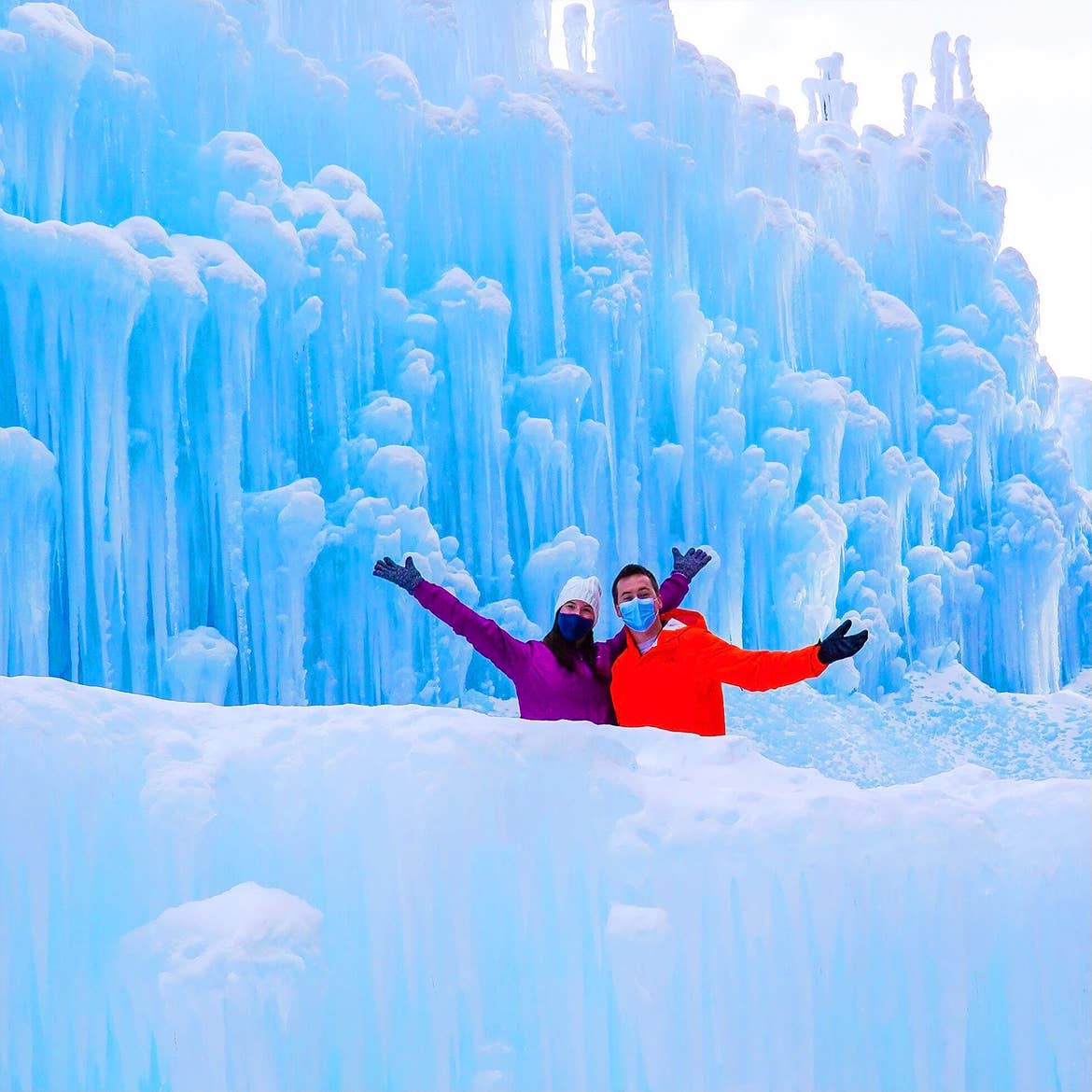 A woman in a white knit hat and purple jacket stands next to a man in an orange jacket in front of an ice castle formation.