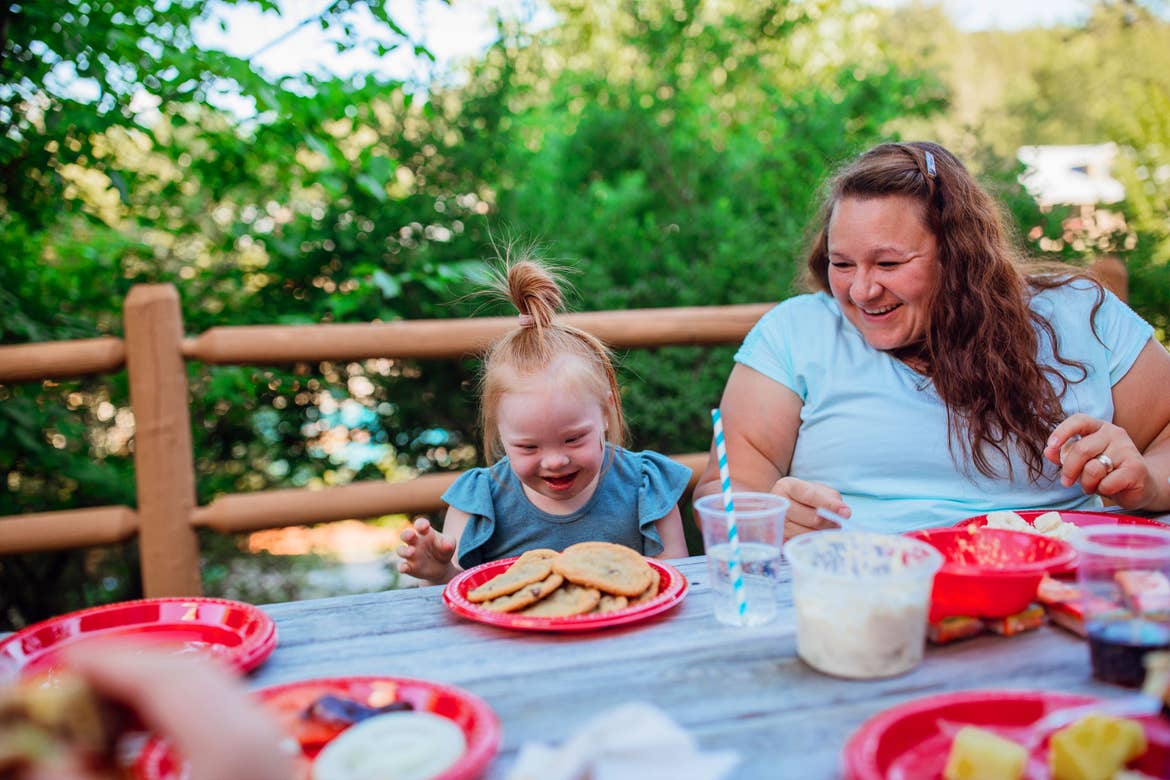 From left to right: A young girl with down syndrome looks at a plate of cookies on a red plate and outdoor table next to a woman.