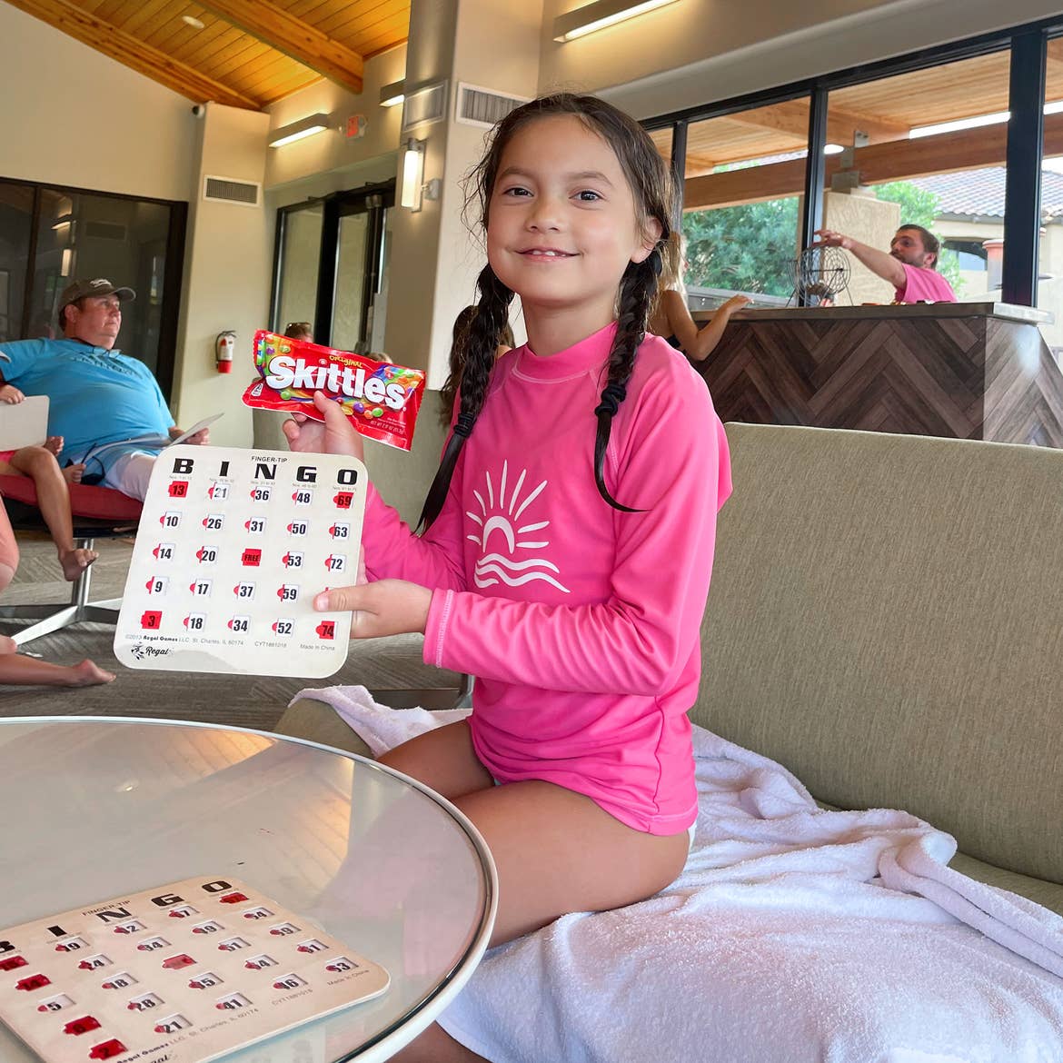 A young girl in a pink rashguard suit holds a complete bingo card and a candy bar while seated near a table.