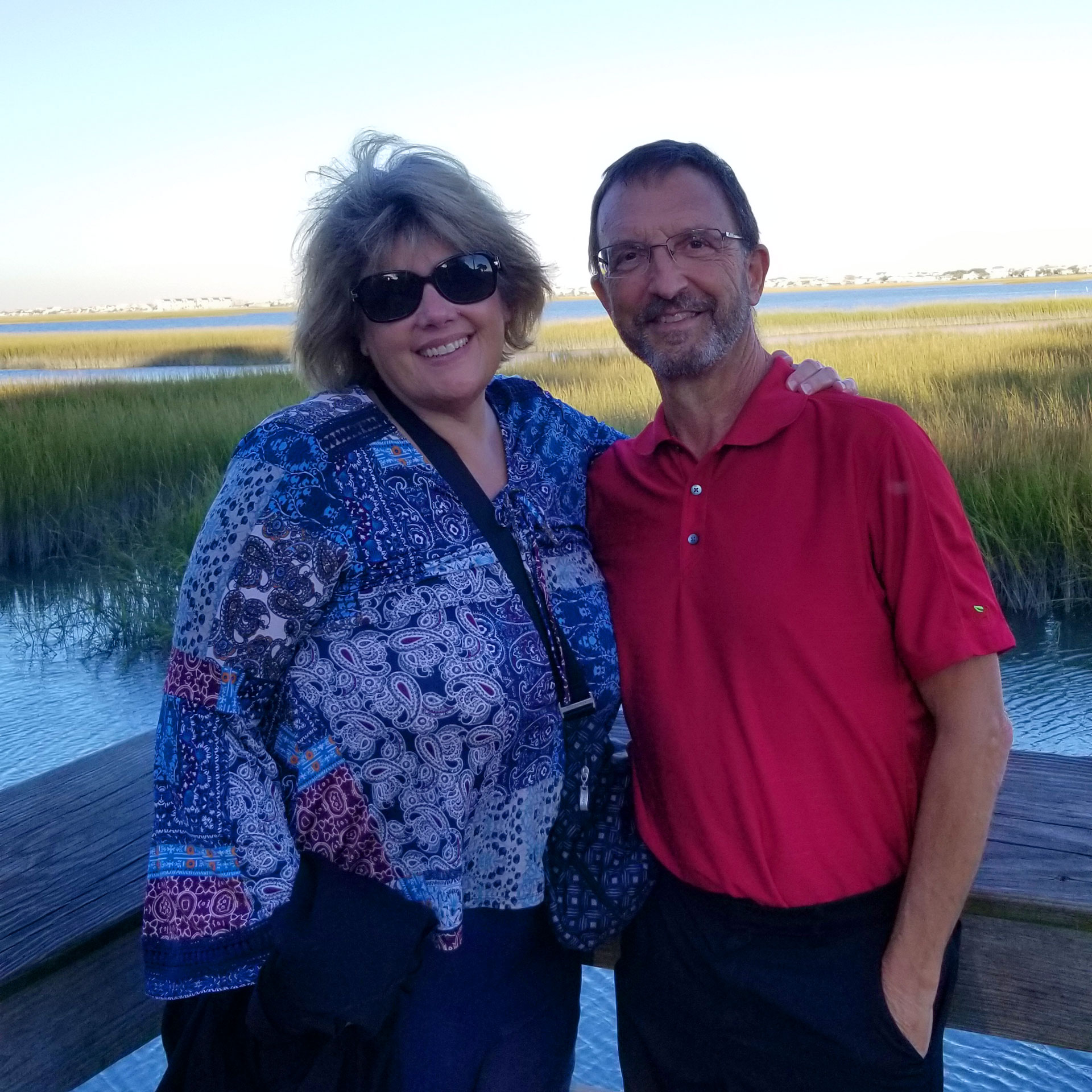 A woman in a floral blouse stands next to a man in a red polo on a wooden platform over a marsh.