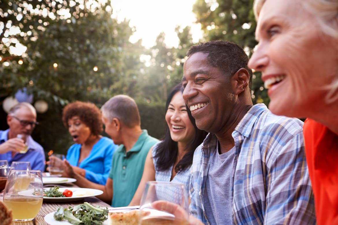Several mature men and women sit at an outdoor picnic table smiling.