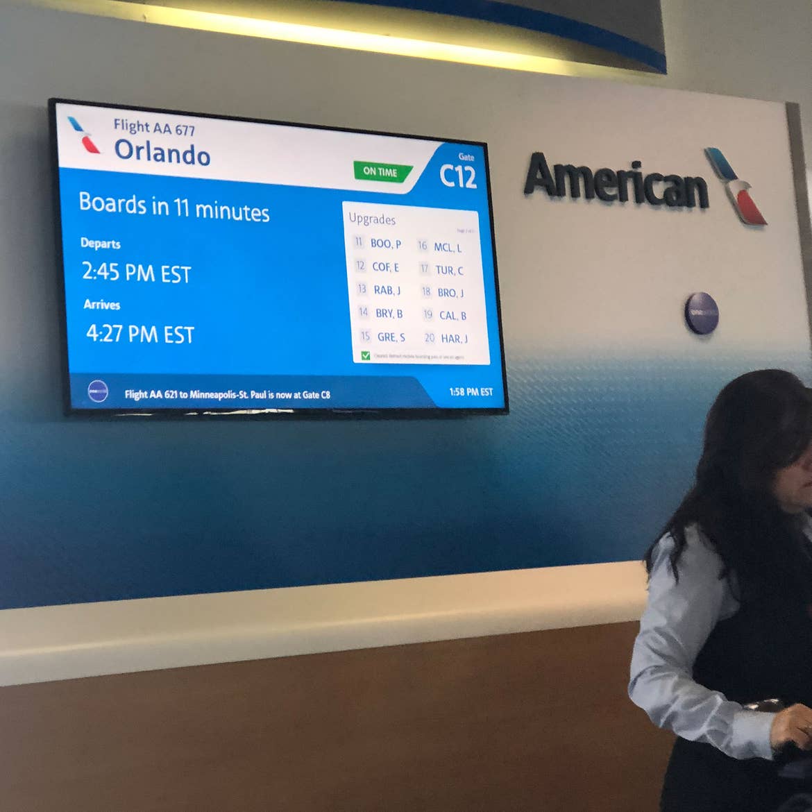 A tv screen with flight gate information as a flight attendant stands nearby.
