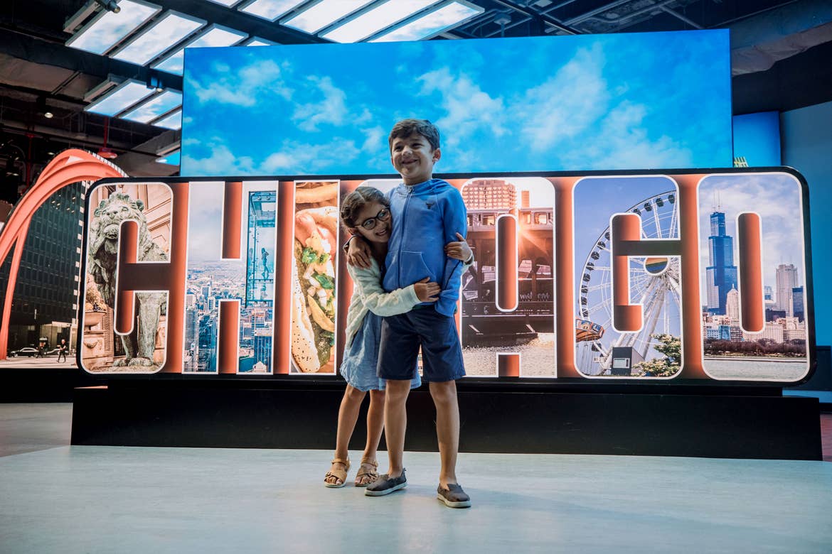 A young girl (left) embraces a young boy (right) in front of a sign that reads, 'Chicago.'