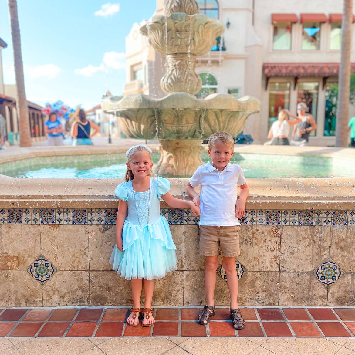 A girl wearing a blue Cinderella dress and a boy wearing a white polo and khaki shorts stand outside near a water fountain.