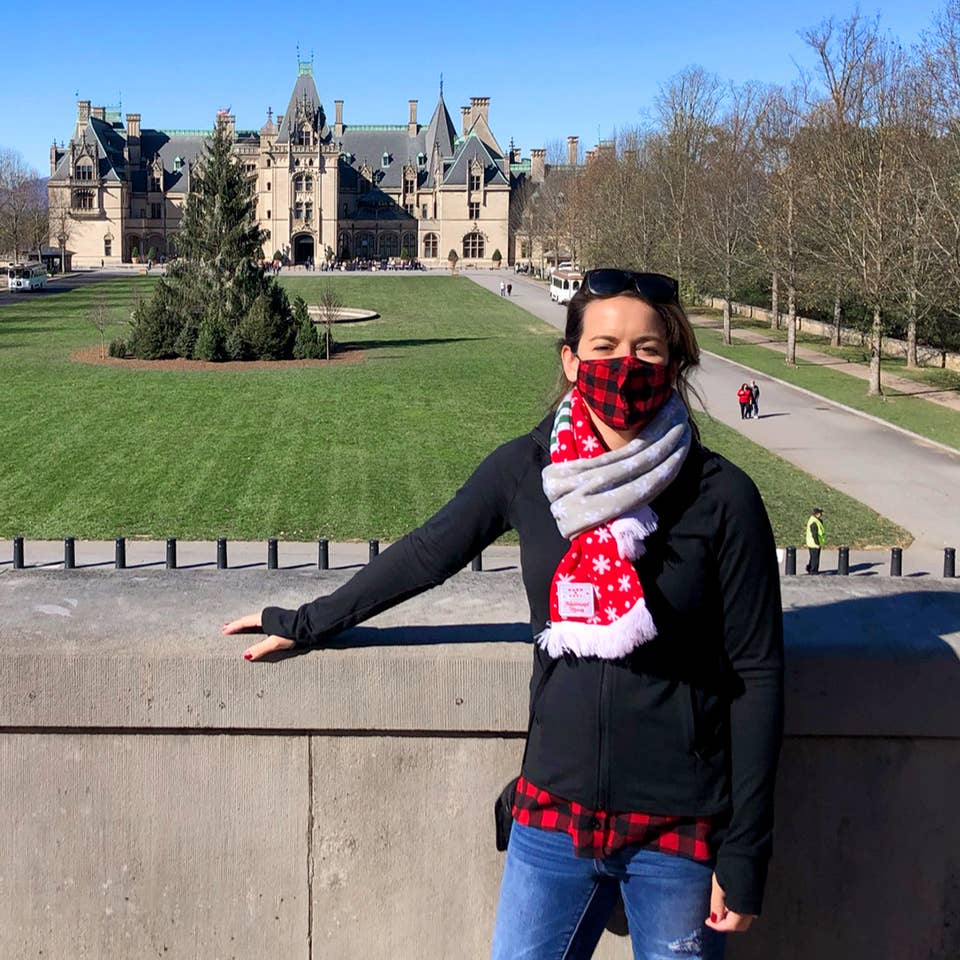 Author, Jenn C. Harmon, stands in front of the Biltmore Estate wearing a buffalo plaid red and black face mask and festive scarf over a black jacket.
