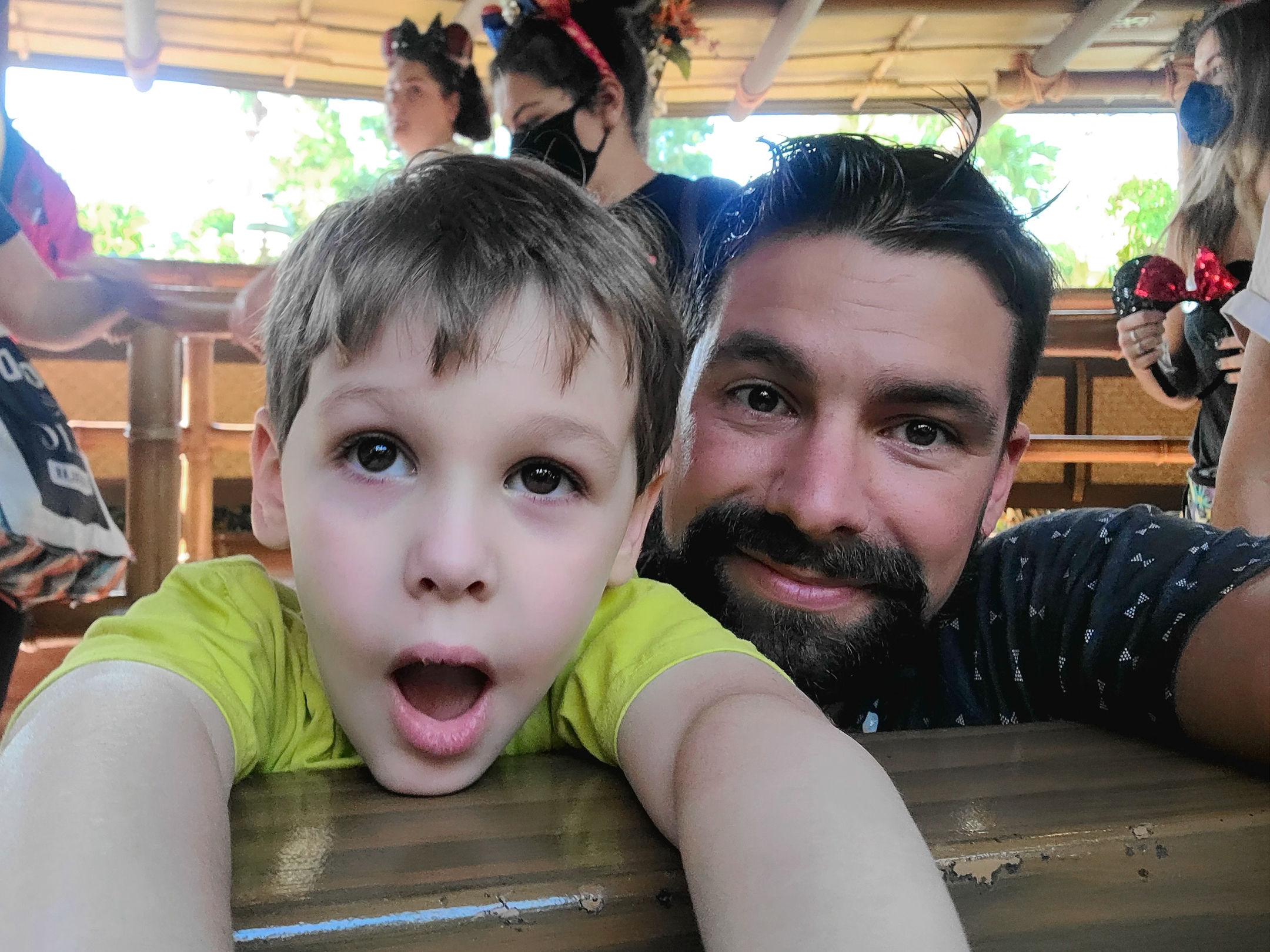 A young boy (left) and man (right) stand in an attraction queue surrounded by guests in safety masks and mouse ears.