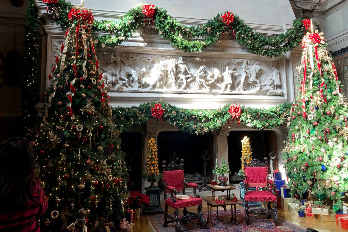 The interior of the Biltmore Estate with an elaborate fireplace mantel decorated with lush Christmas garlands and surrounded by red seating and Christmas trees.