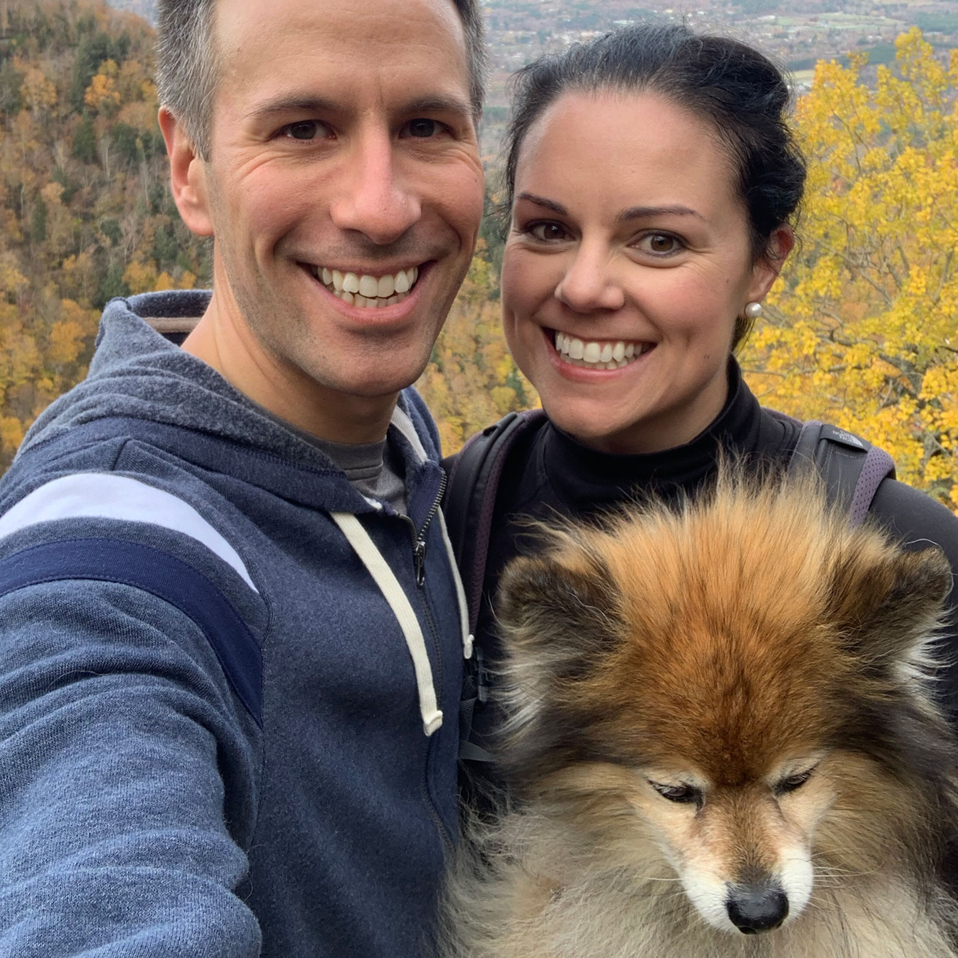 Authors, Lauren Layne and Anthony LeDonne, stand in front of fall foliage with Bailey the Pomeranian.
