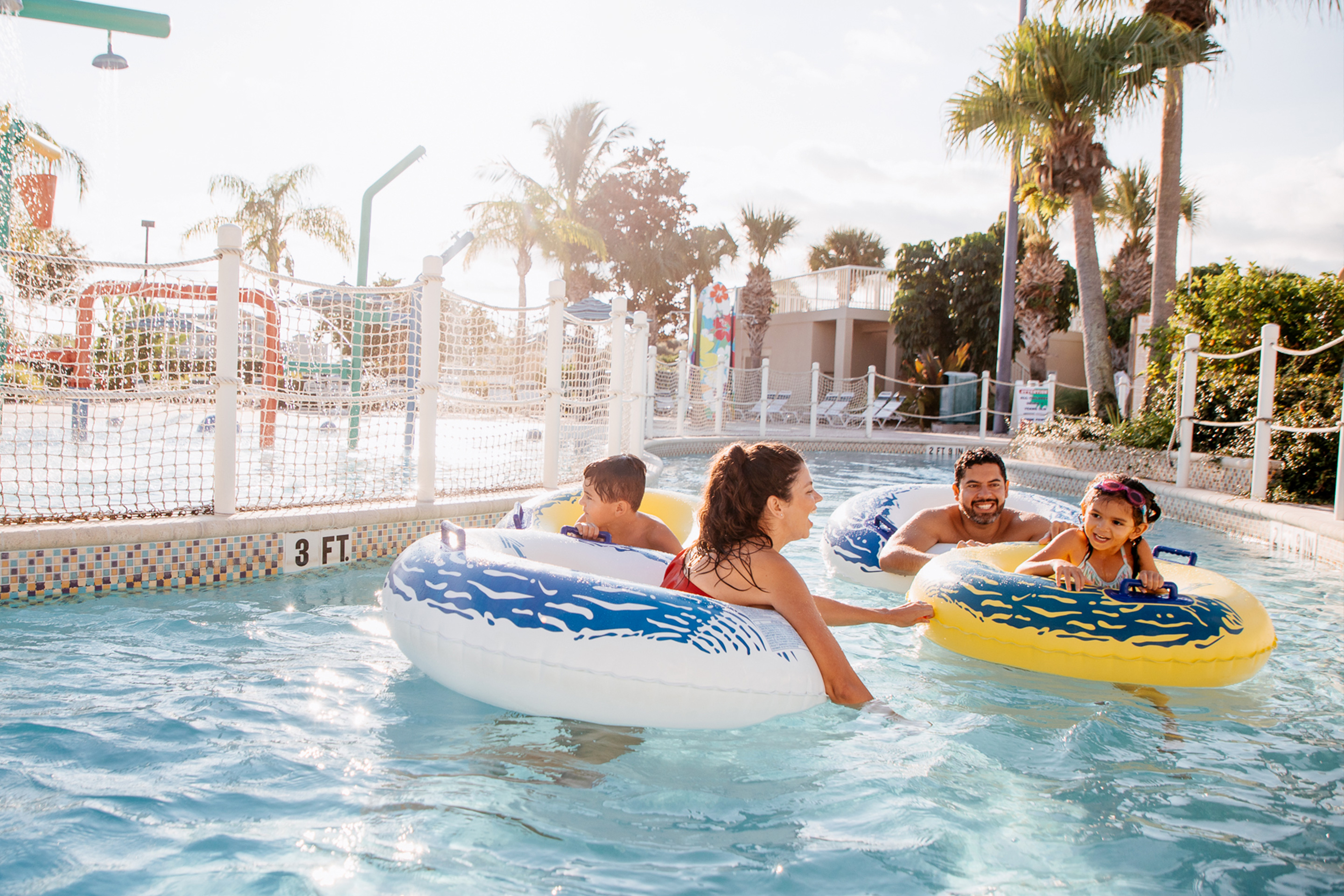 A man, woman, and young boy and girl wear swimsuits while floating in yellow inner tubes in an outdoor lazy river.