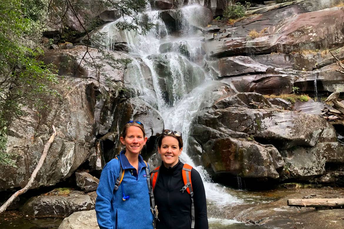 Featured Contributor, Jennifer C. Harmon (right) and her friend pose together in front of a waterfall wearing backpacks and jackets.