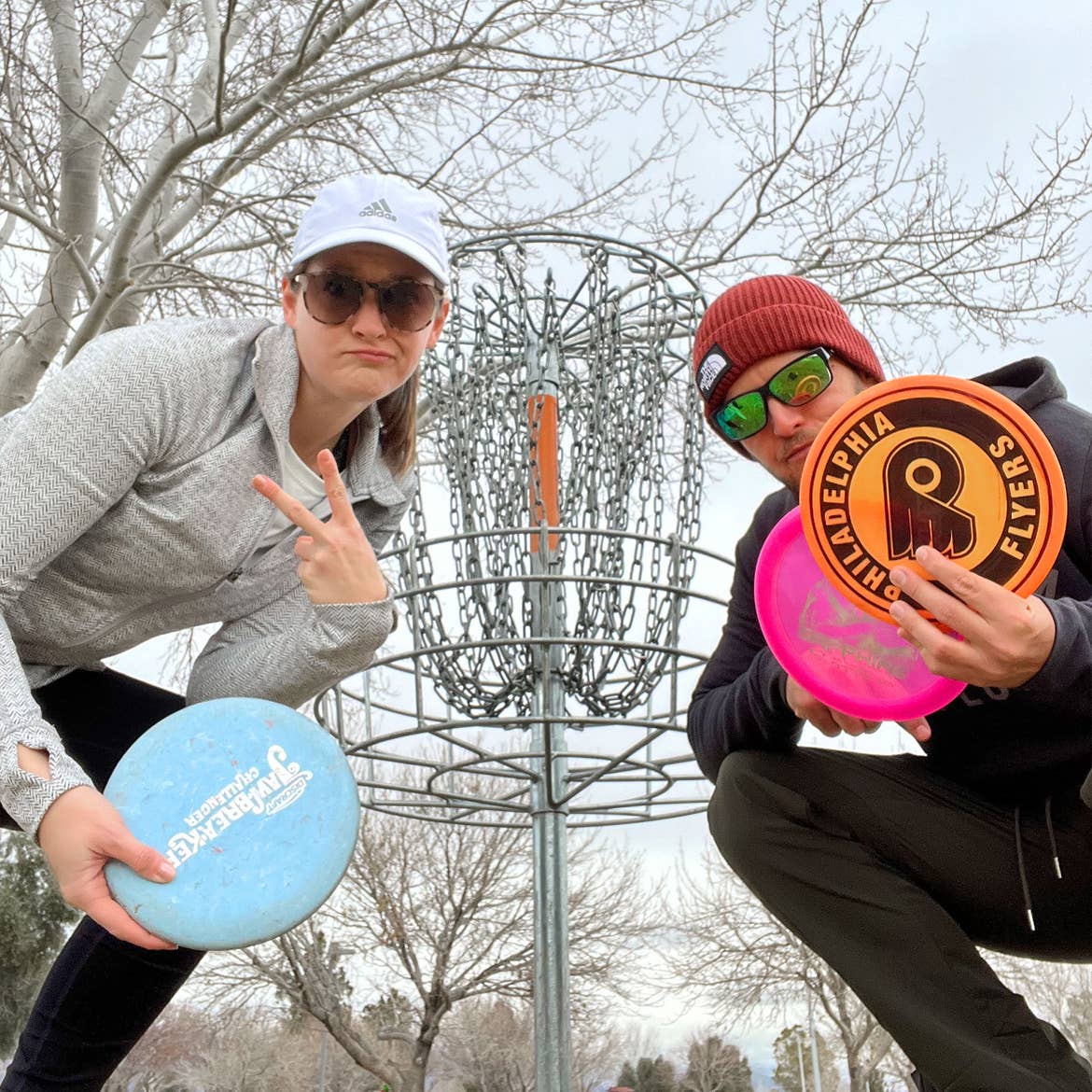 A woman wearing a grey pullover, white cap and holding a blue frisbee disc stands next to a man in a black pullover and red knitted cap holding two frisbee discs near an outdoor disc golf basket.