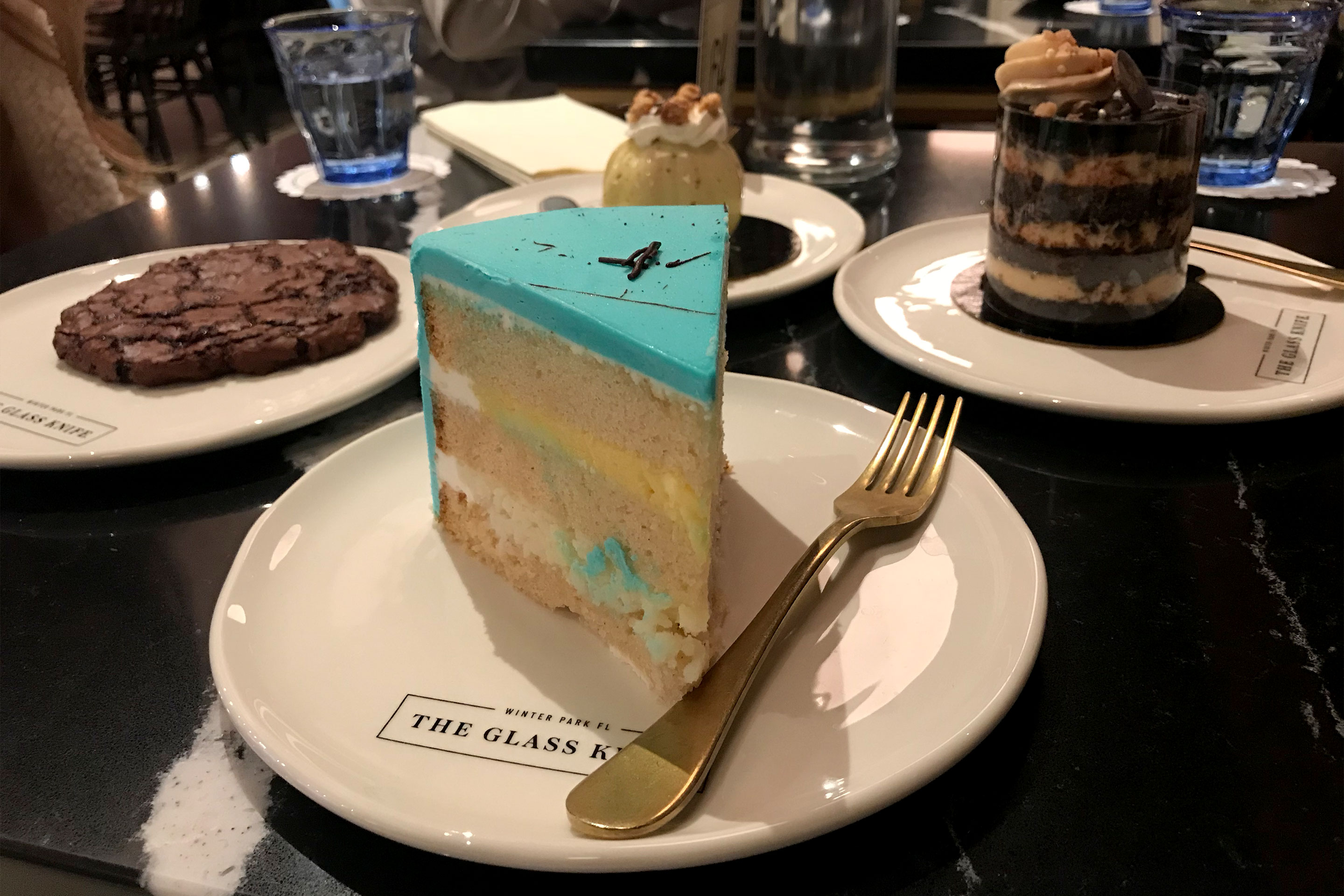 Several plates containing different desserts like cookies, blue cake, mousse are placed on a table.