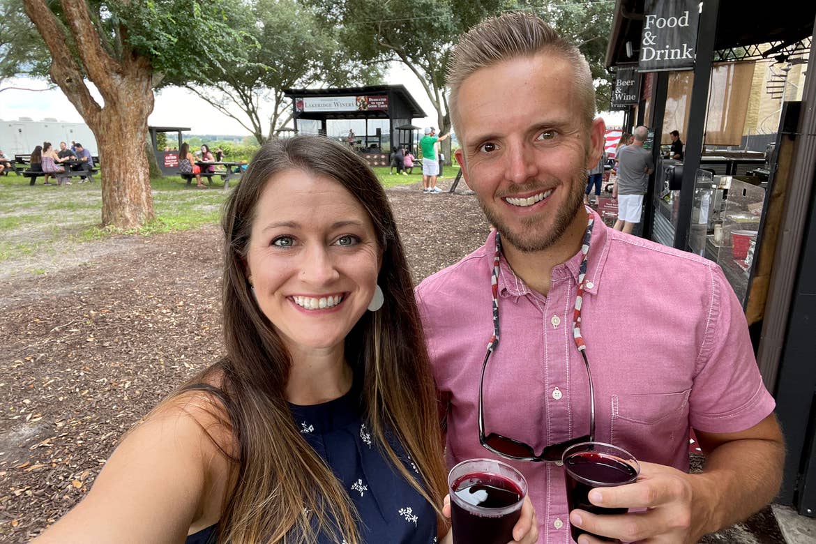 A man (right) wears a red button-up shirt next to a woman (left) wearing a navy dress while holding wine near winery vendor stalls outdoors.