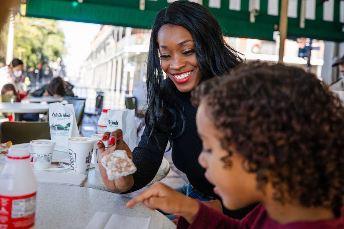 Featured contributor, Sally Butan (left) wears a black shirt holding a beignet while her son (right) wears a maroon shirt under a green canopy at Cafe Du Monde eating a powdery beignet.