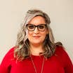 Misty Treadway wears a red blouse and black glasses in front of a beige wall.