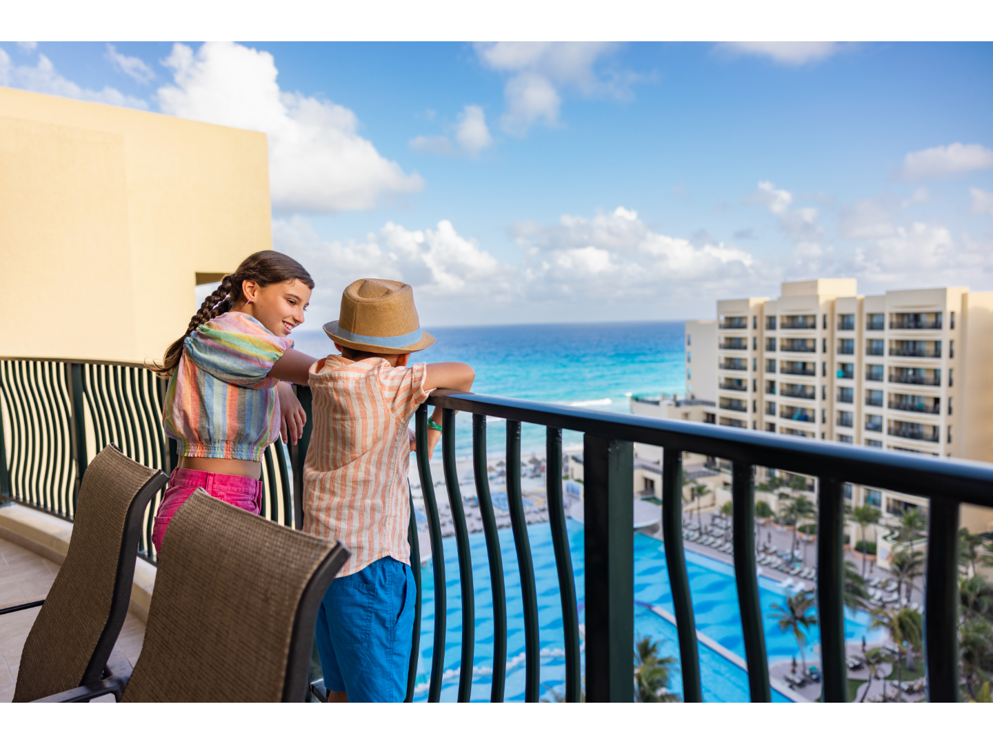 Two kids enjoying a balcony view overlooking a resort pool, tall hotel buildings, and the turquoise ocean.