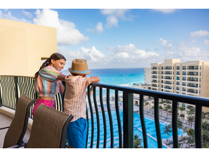 Two kids enjoying a balcony view overlooking a resort pool, tall hotel buildings, and the turquoise ocean.