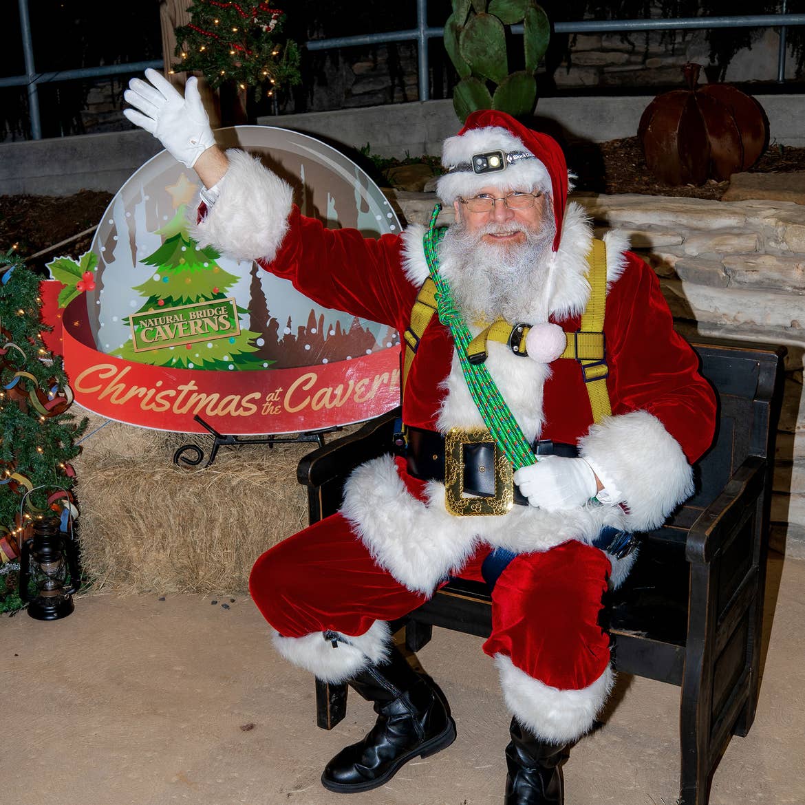 Santa 'Spelunker' Clause sits wearing his red suit, and climbing harness while waving to guests from a black bench in front of a snow globe that reads, 'Christmas & Caverns.'