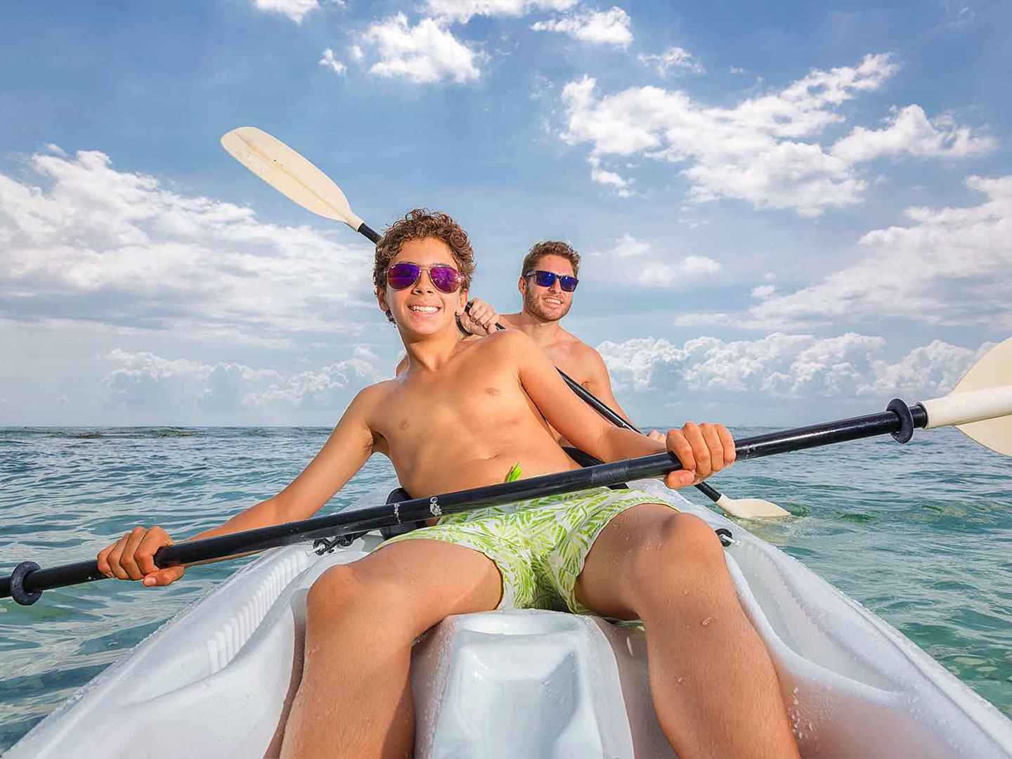Father and son, smiling and wearing sunglasses while kayaking on the ocean at The Royal Haciendas in Playa del Carmen, Mexico
