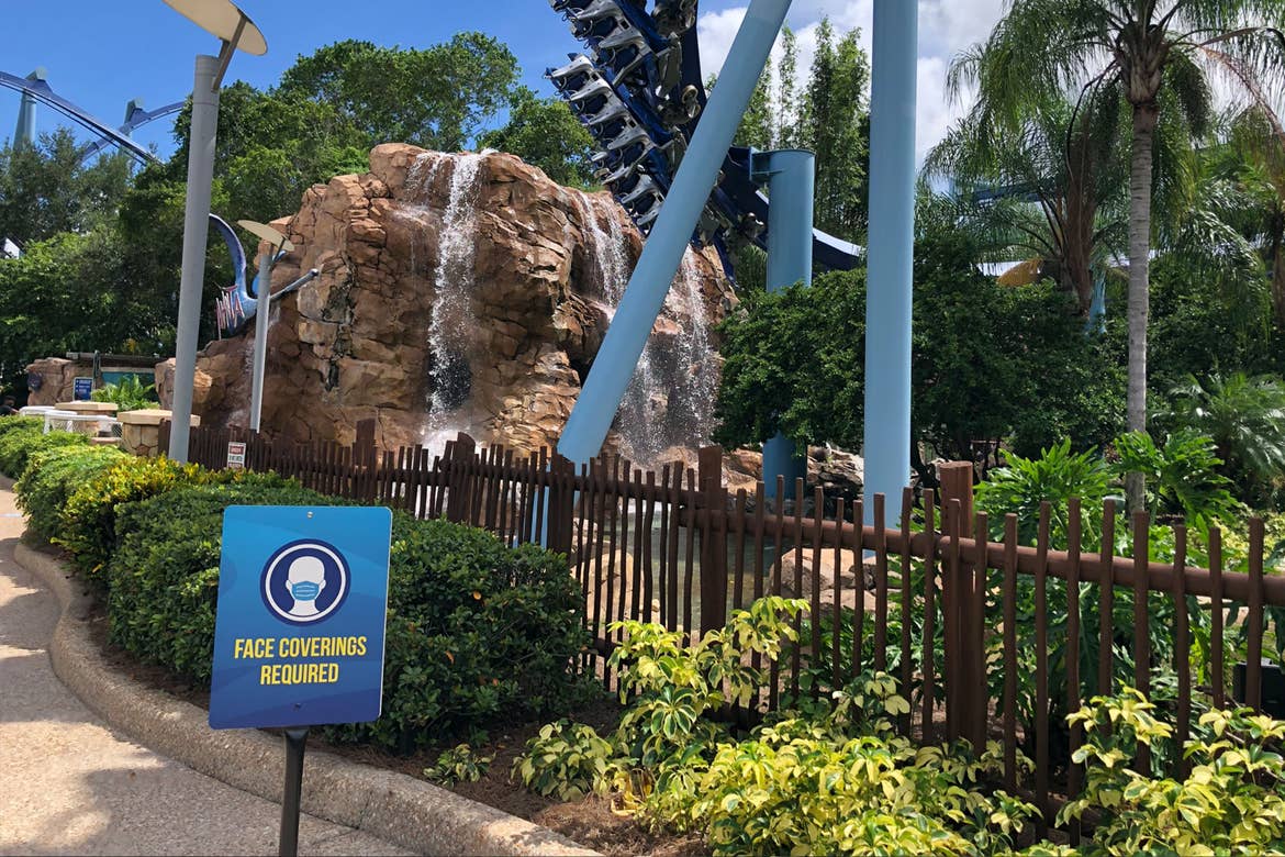 A sign indicating to guests that 'Face Masks Are Required' stands in front of a rollercoaster to maintain social distancing guidelines.