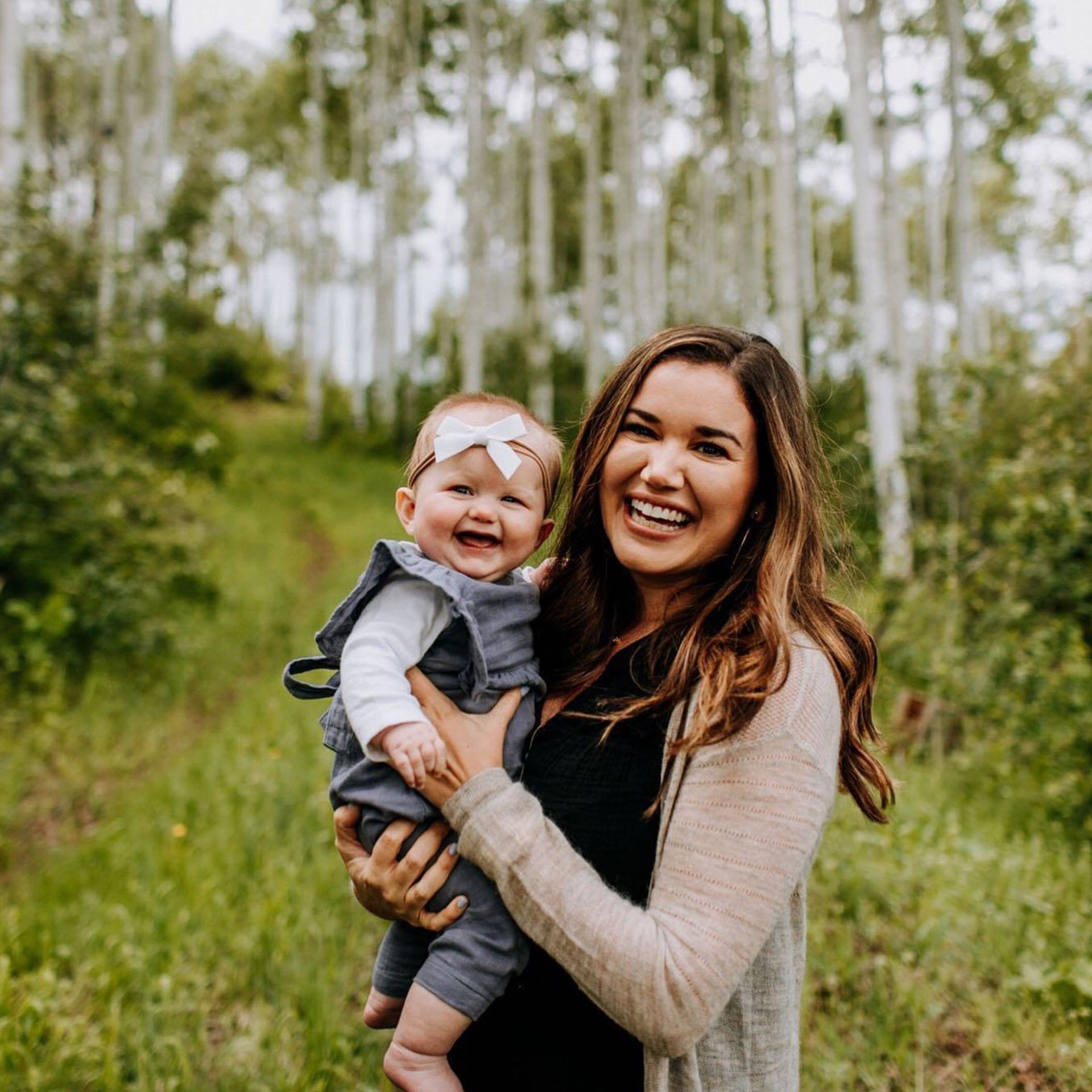 A mother holds her infant daughter in front of a grassy path and birch trees.