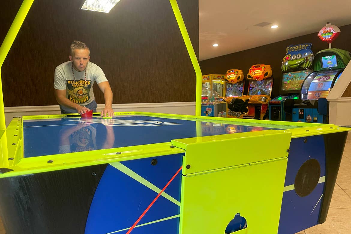 A man in a grey t-shirt stands at a neon-colored air hockey table in an indoor arcade.