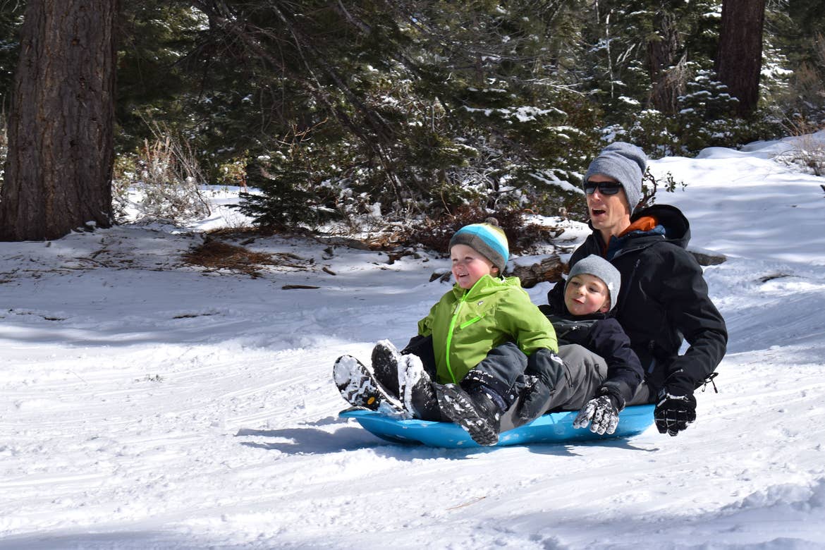A man and two children sit on a black sled going down a snowy hill.
