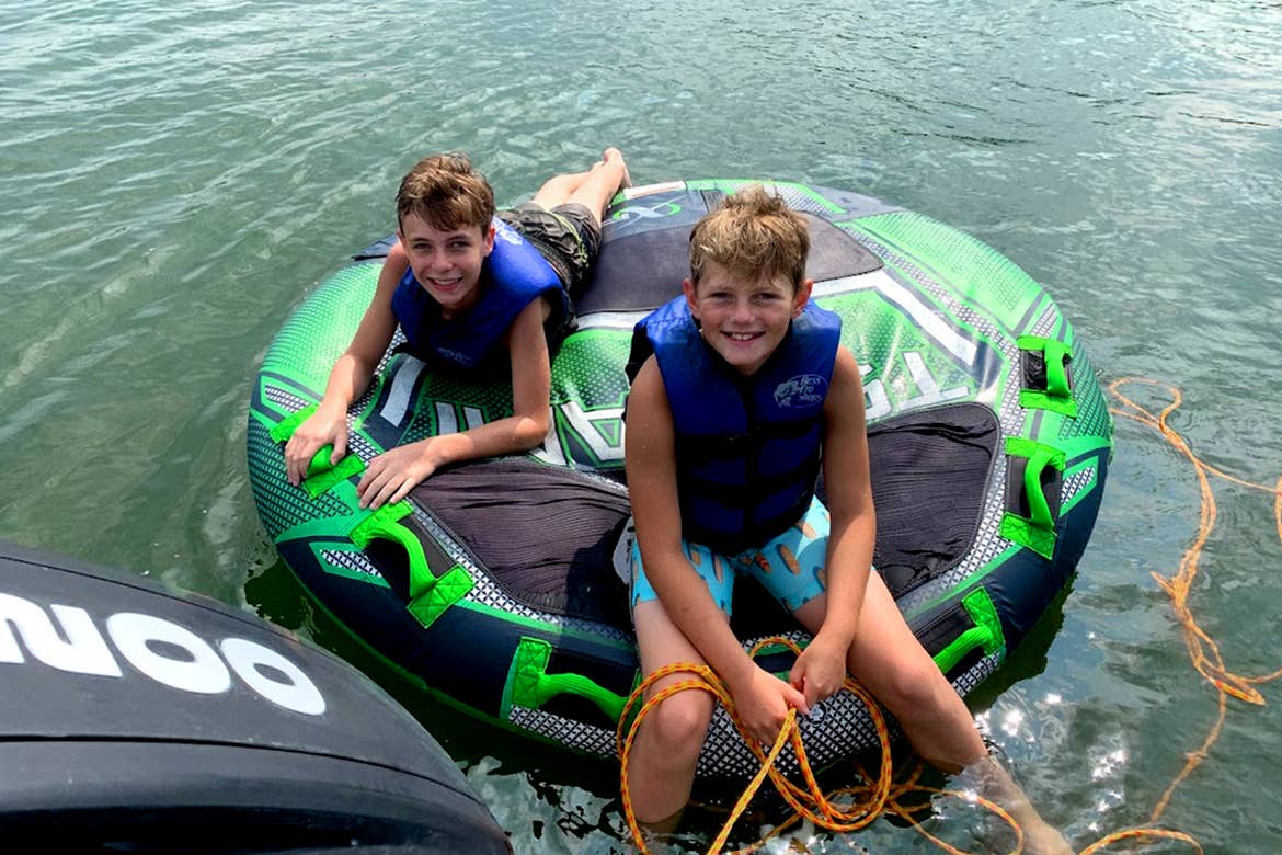 Two kids with lifejackets sit on a tube being towed by boat.