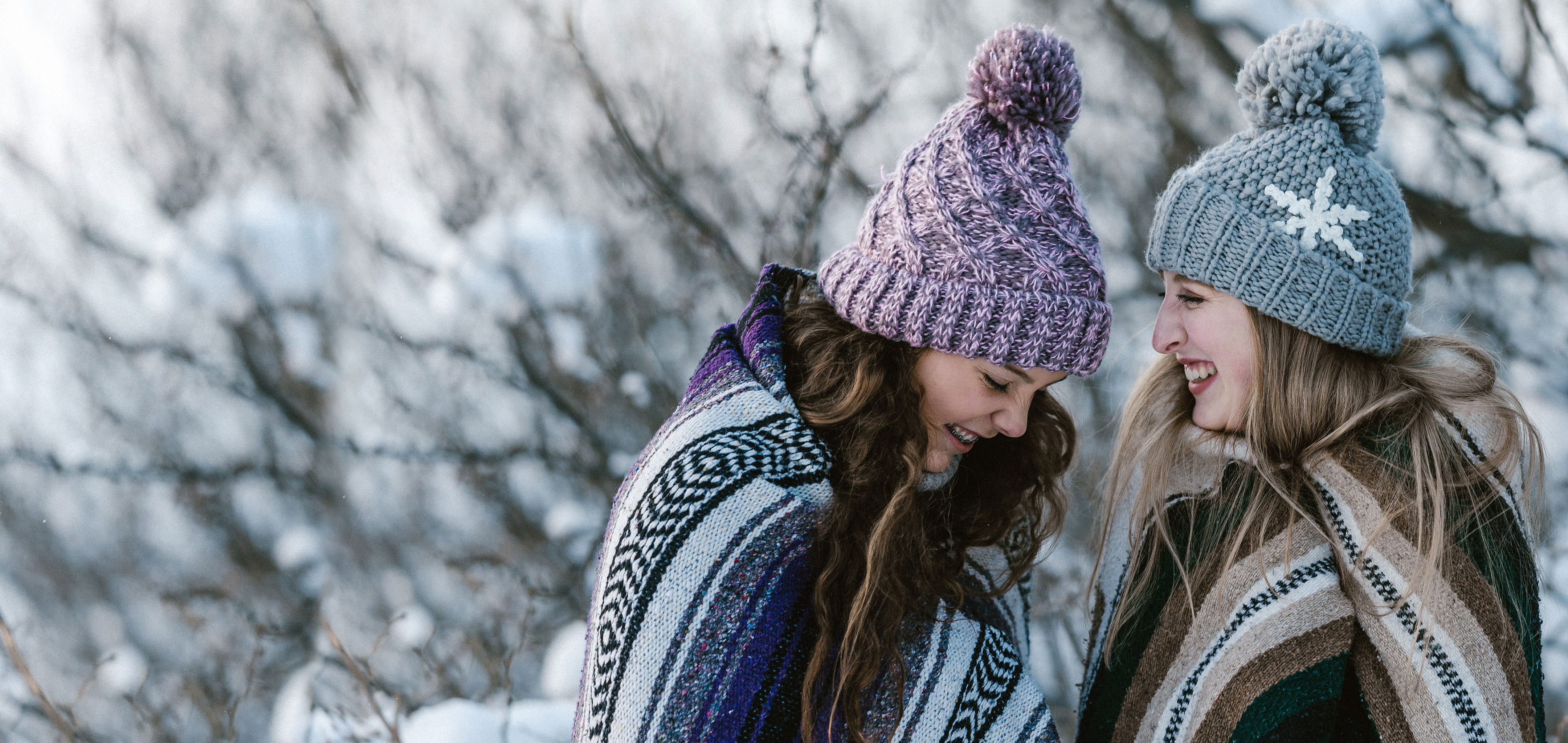 Two girls outside in the snow wearing blankets and beanies while laughing.
