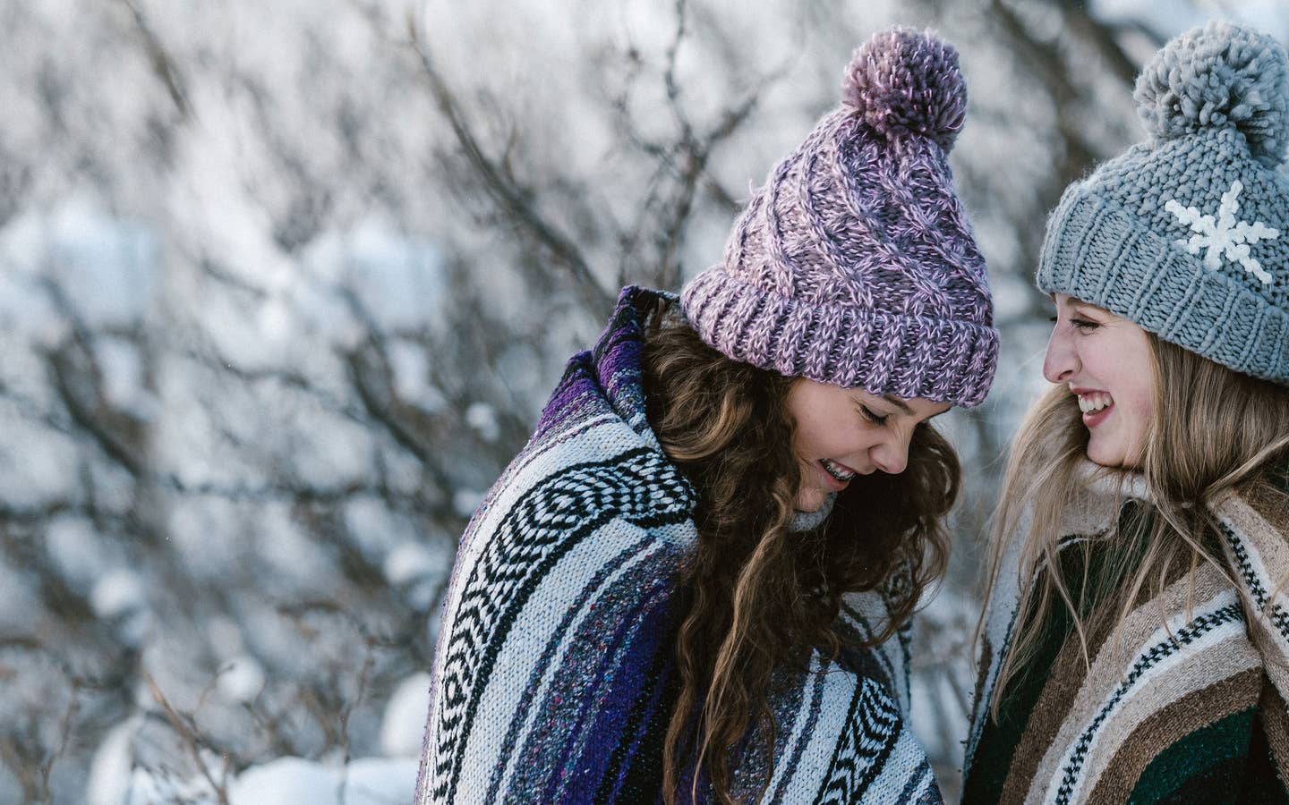 Two girls outside in the snow wearing blankets and beanies while laughing.