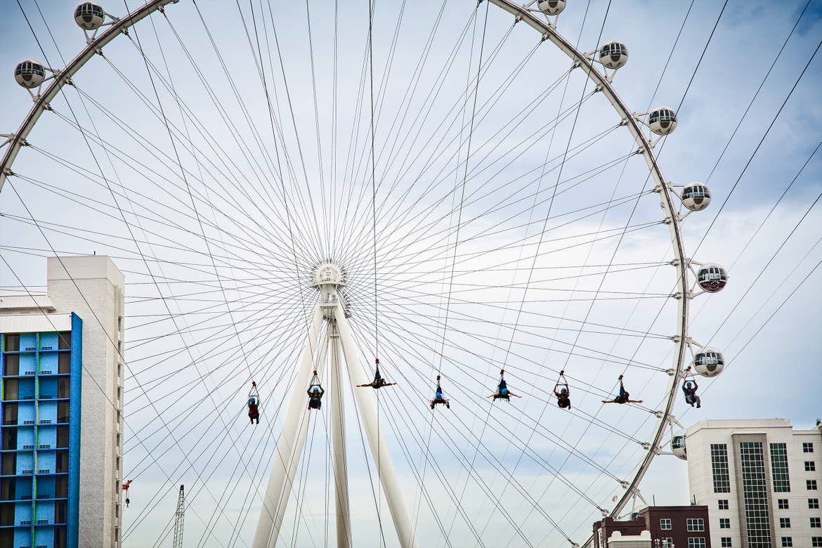 Several people are harnessed onto ziplines as they approach the 'High Roller' ferris wheel under a cloudy sky.