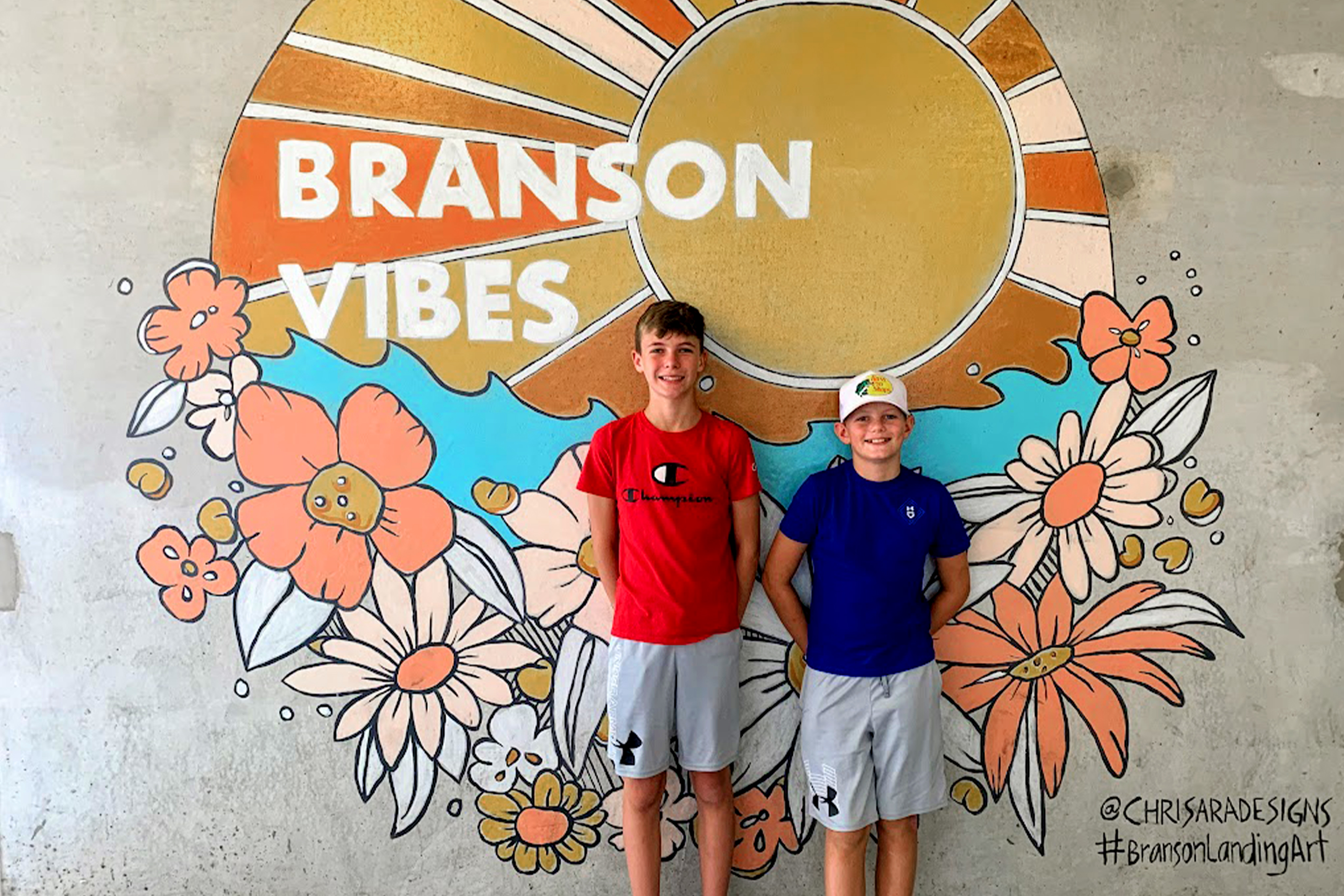 Two boys stand in front of a mural that reads,  'Branson Vibes'.