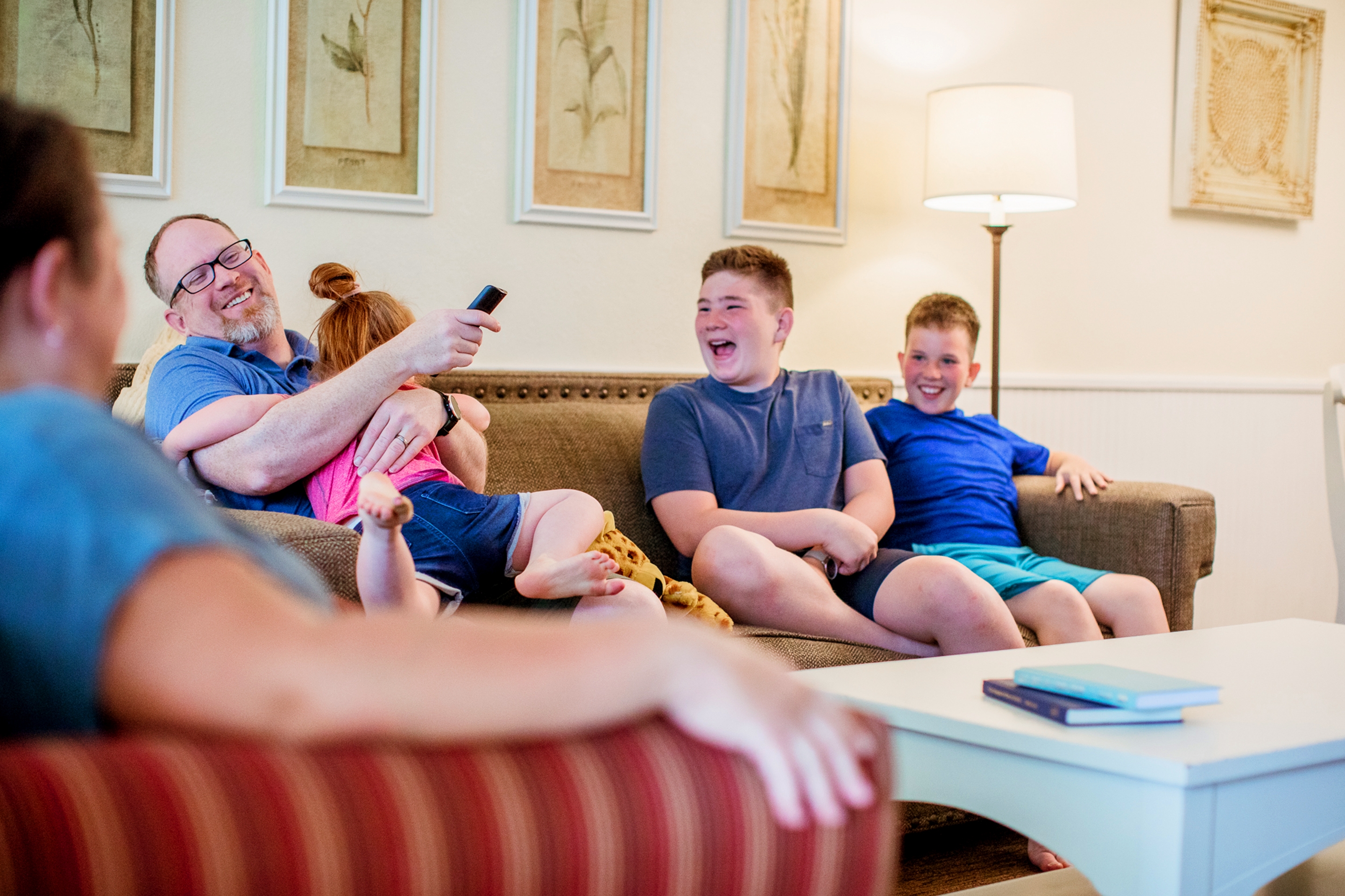 A caucasian family of five (left to right: A woman, a man, a girl, and two tween boys) sit on a couch as the man holds a remote and a young girl inside a villa.
