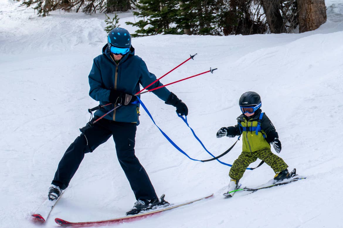 A man in blue winter apparel skis next to a young boy being trained on skis with a harness on a snowy slope.
