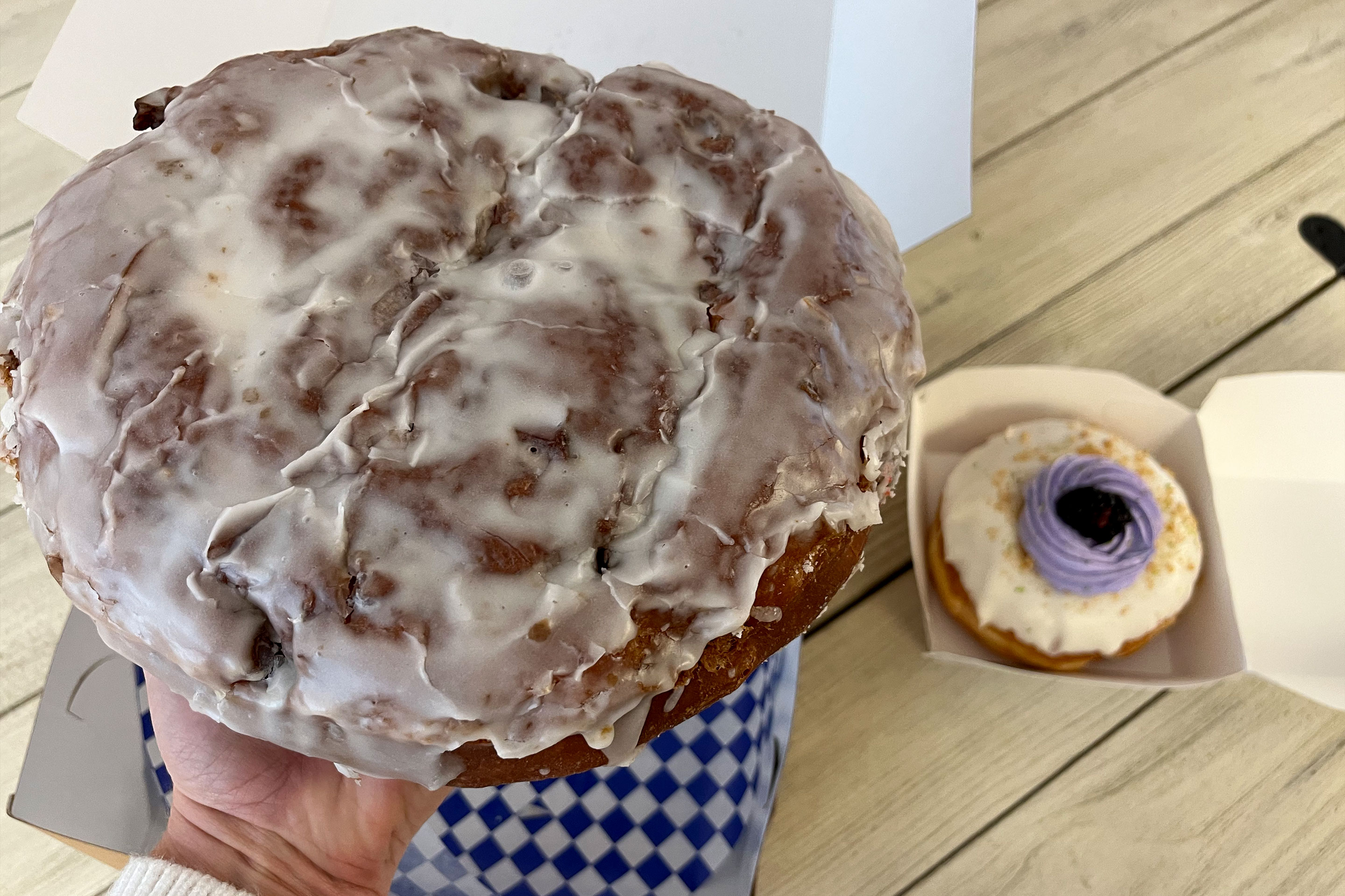 A woman holds a large cinnamon roll over a takeout box with blue and white checkered paper near another takeout box with a doughnut covered in white and blue frosting.