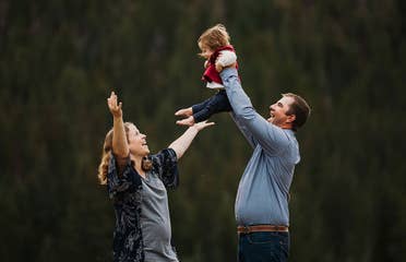 A mother (left) lifts her expressive arms as a father (right) holds up their child in front of a wooded area.