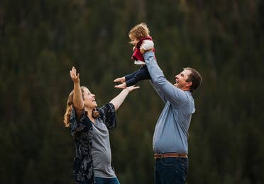 A mother (left) lifts her expressive arms as a father (right) holds up their child in front of a wooded area.