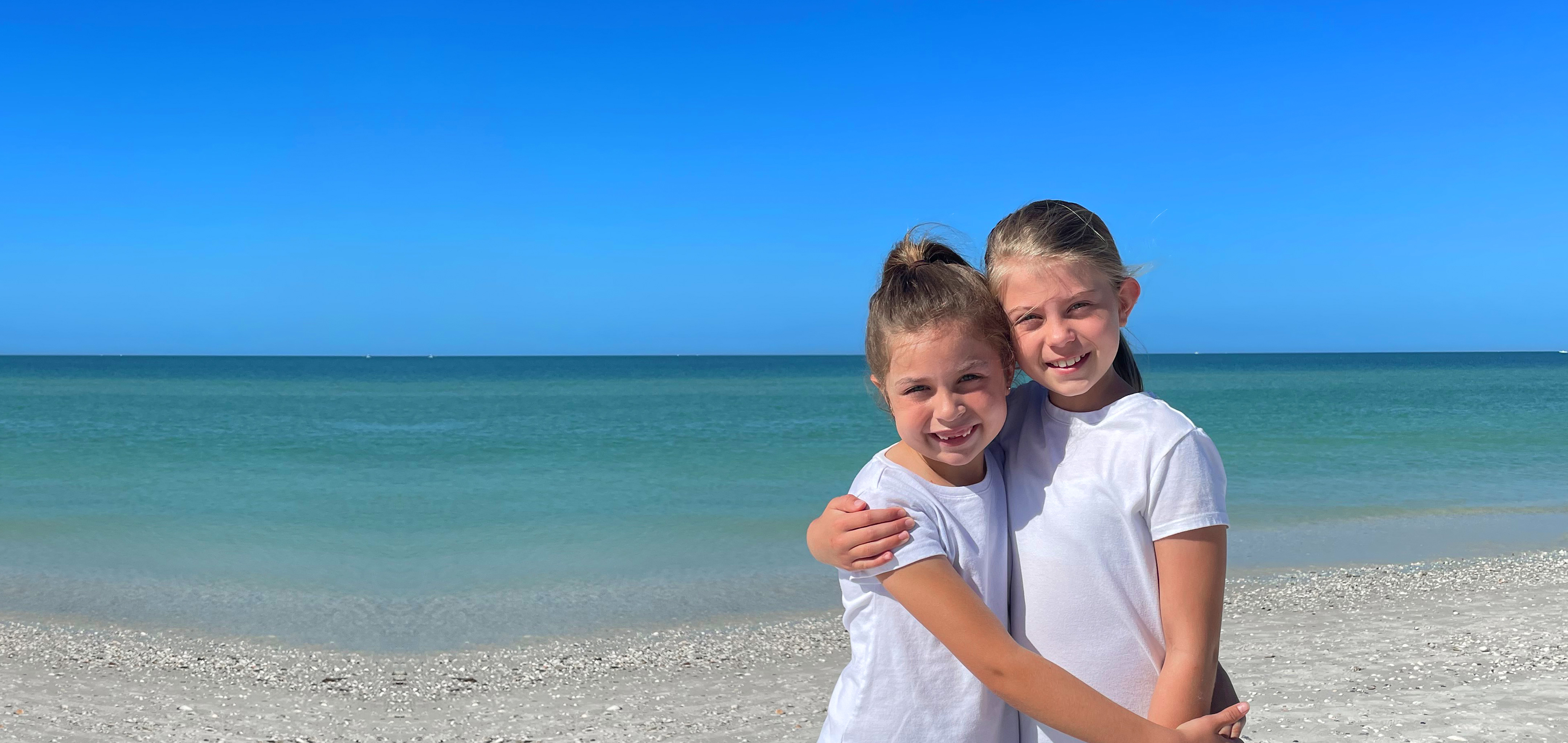 Two caucasian girls wearing white t-shirts and running shorts stand on a beach.