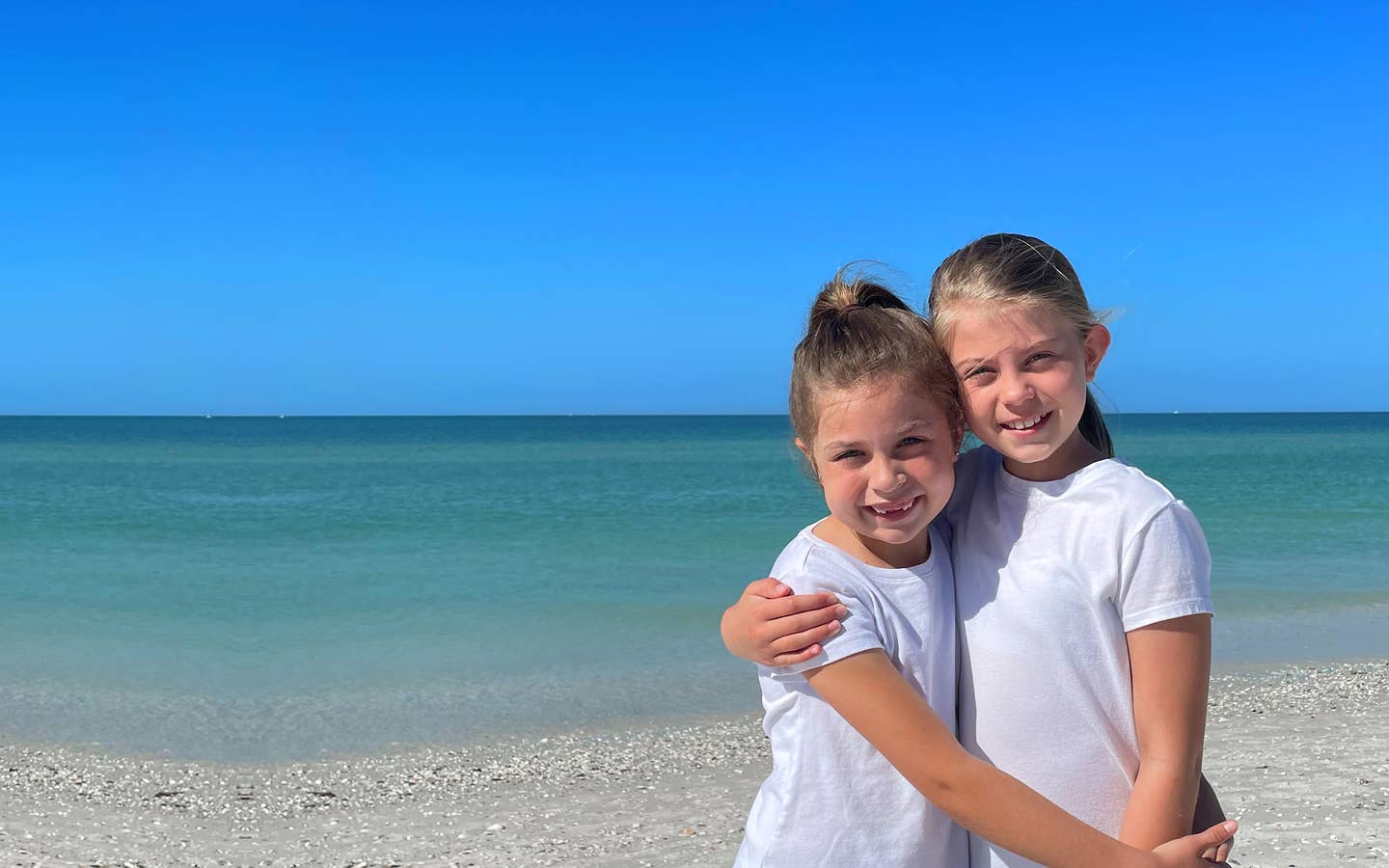 Two caucasian girls wearing white t-shirts and running shorts stand on a beach.