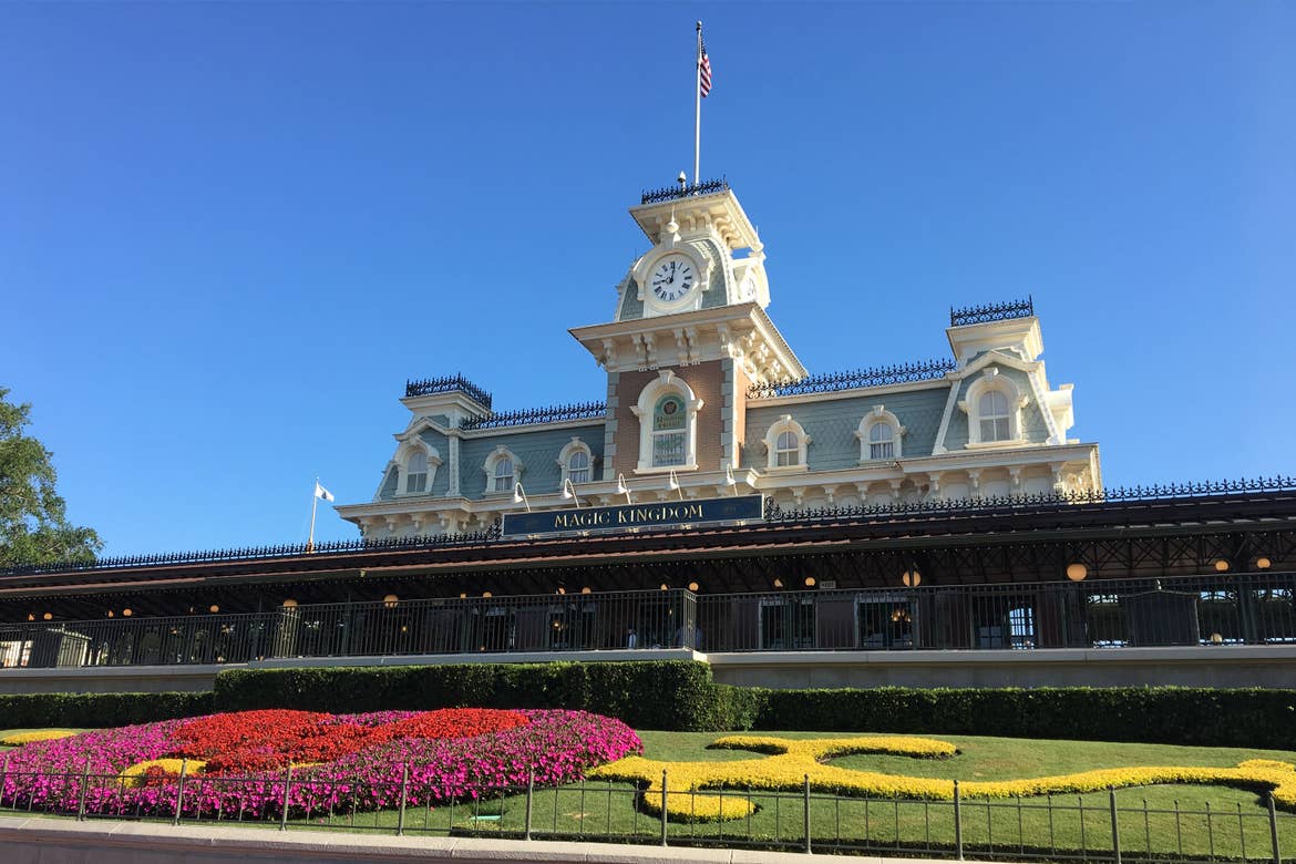 The exterior of the Walt Disney World Railroad - Main Street, U.S.A. Train Station under a blue sky and surrounded by colorful flowers arranged in the shape of Mickey Mouse.