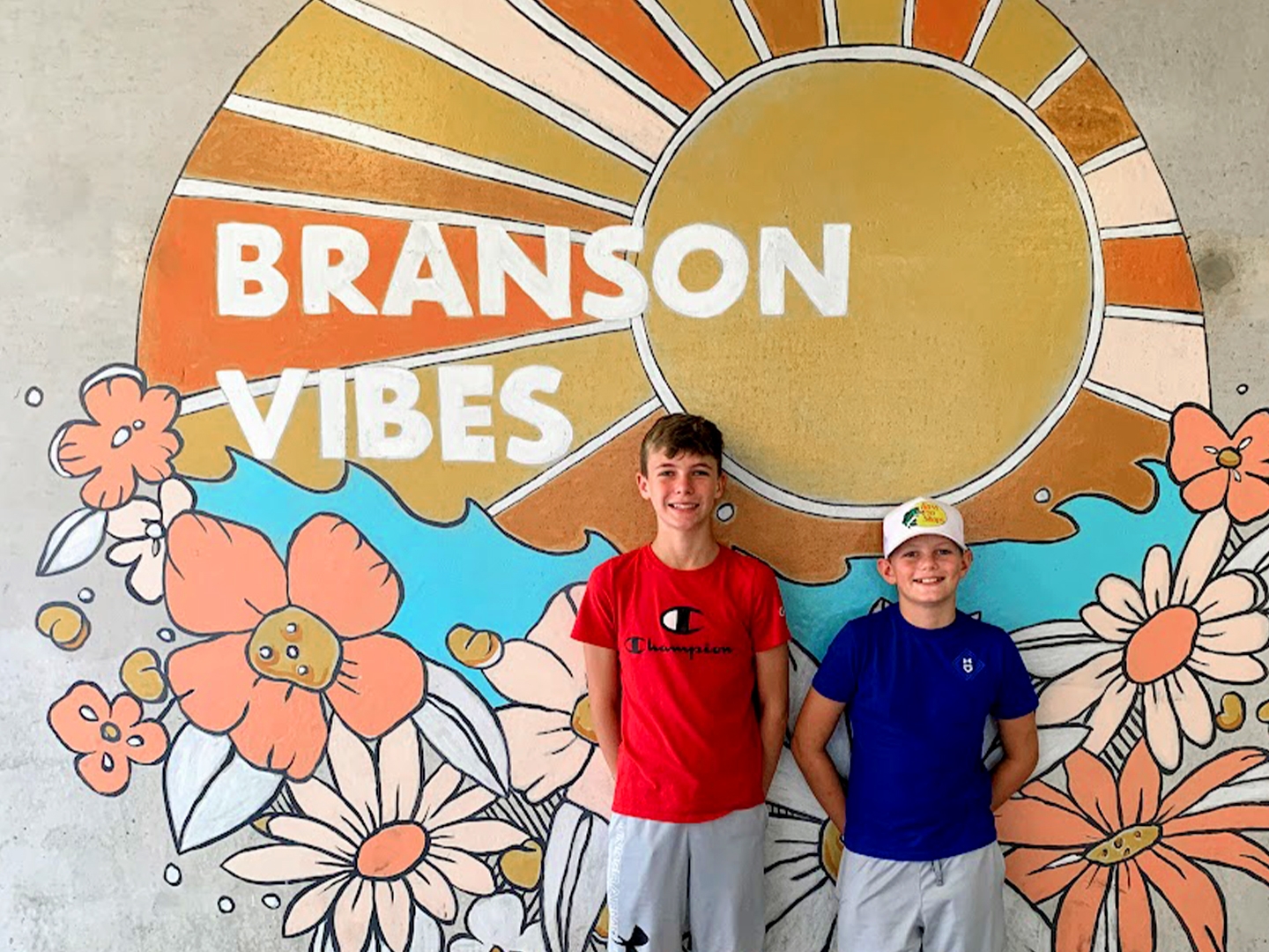 Two boys stand in front of a mural that reads,  'Branson Vibes'.