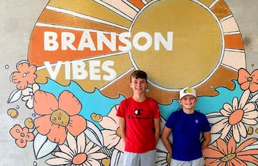 Two boys stand in front of a mural that reads, 'Branson Vibes'.