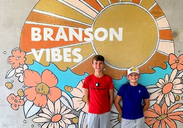 Two boys stand in front of a mural that reads,  'Branson Vibes'.