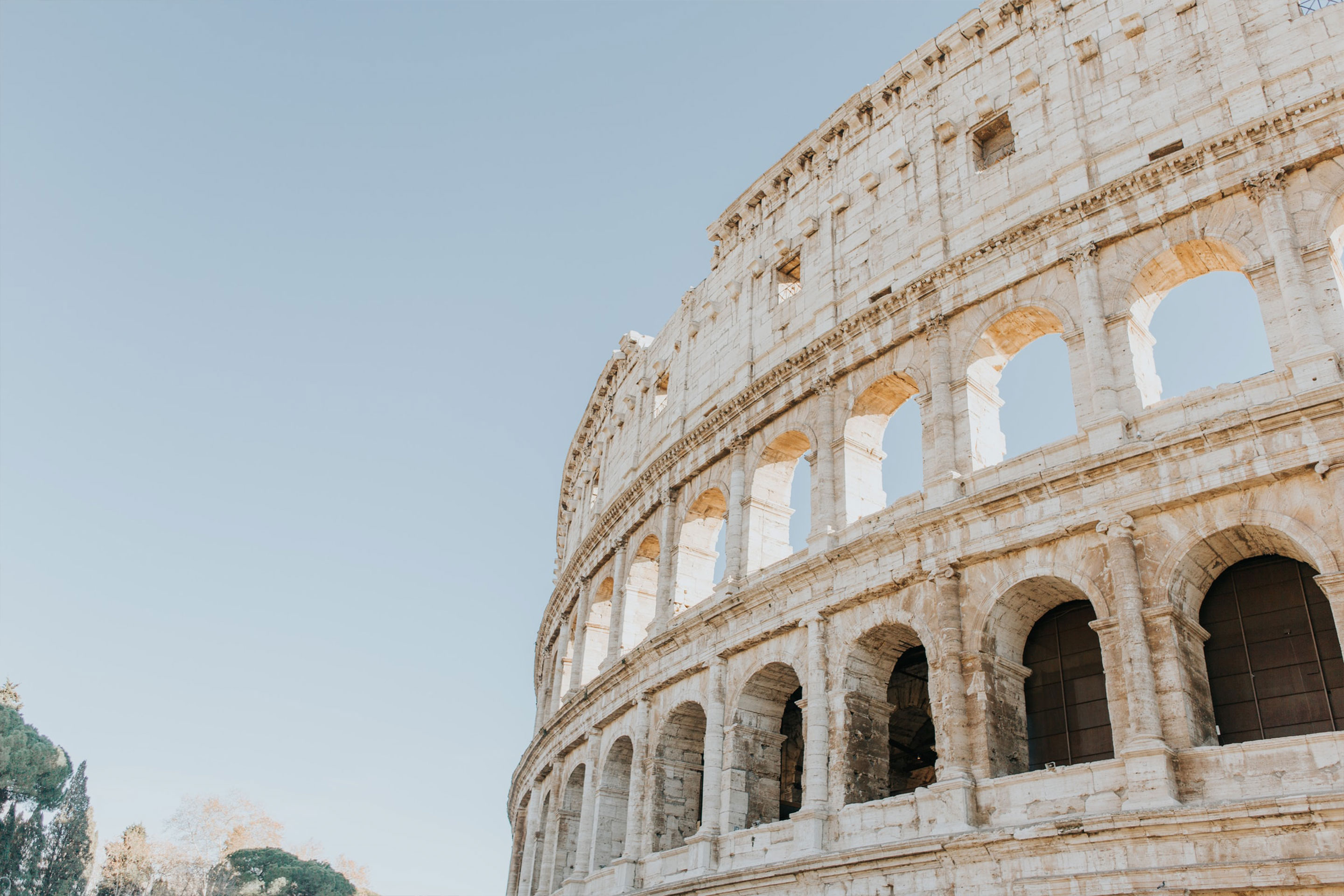 The white stone exterior of the Coliseum in Rome, Italy under a light blue sky.