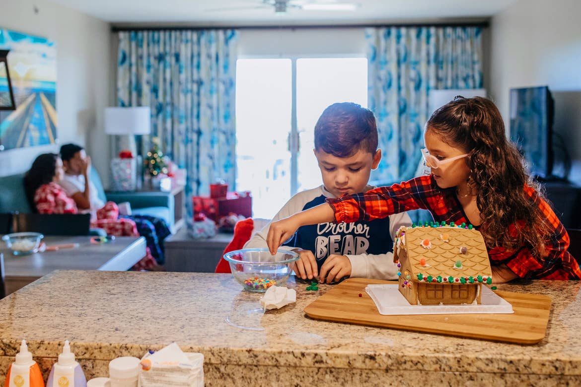A young boy and girl build a gingerbread house as the parents sit in the background.